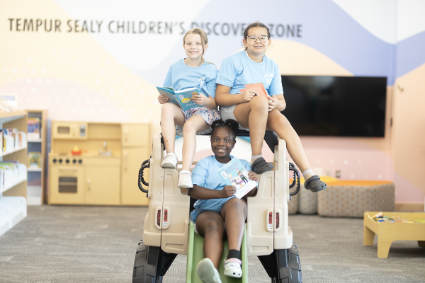 Three children sit together on a toy car at the Marksbury Family Branch.